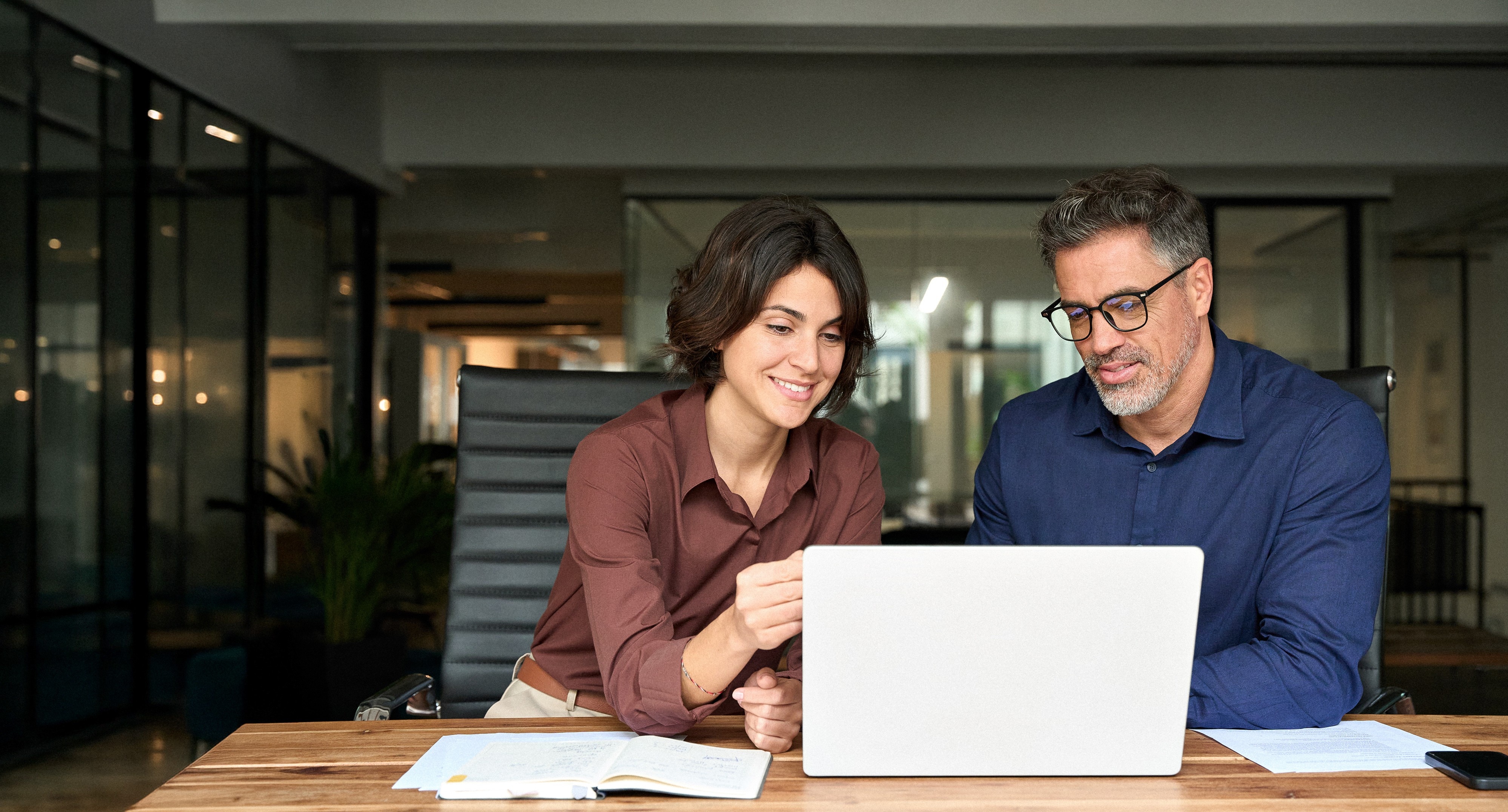 Female executive manager explaining corporate project plan to male colleague sitting at office desk. Business team of two busy professionals working together discussing ai solutions using laptop.