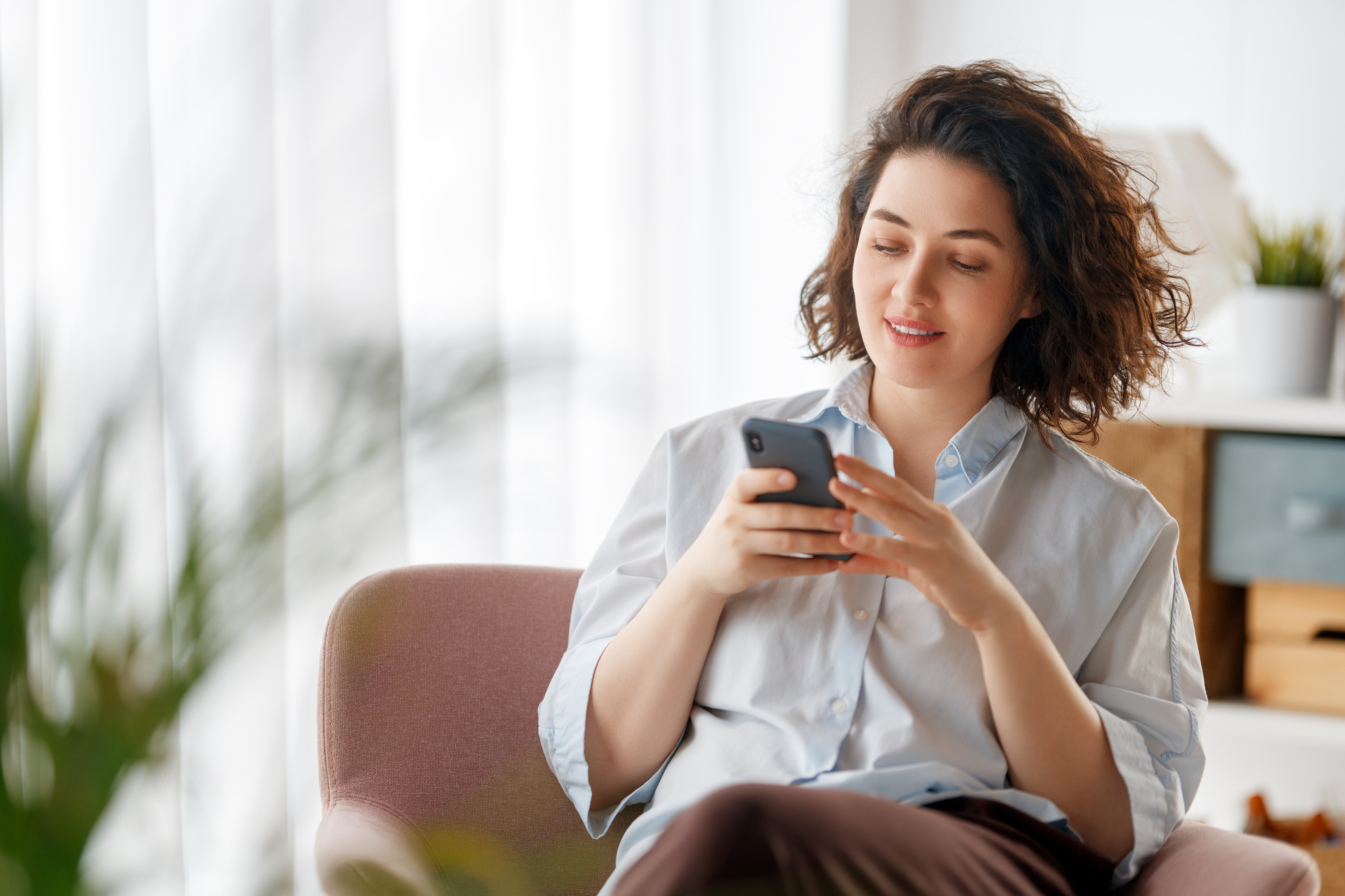 Happy casual beautiful woman is using a phone sitting on armchair at home.