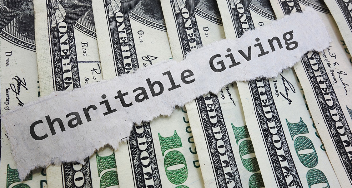 alt=Stack of $100 bills lying under strip of paper labeled Charitable Giving