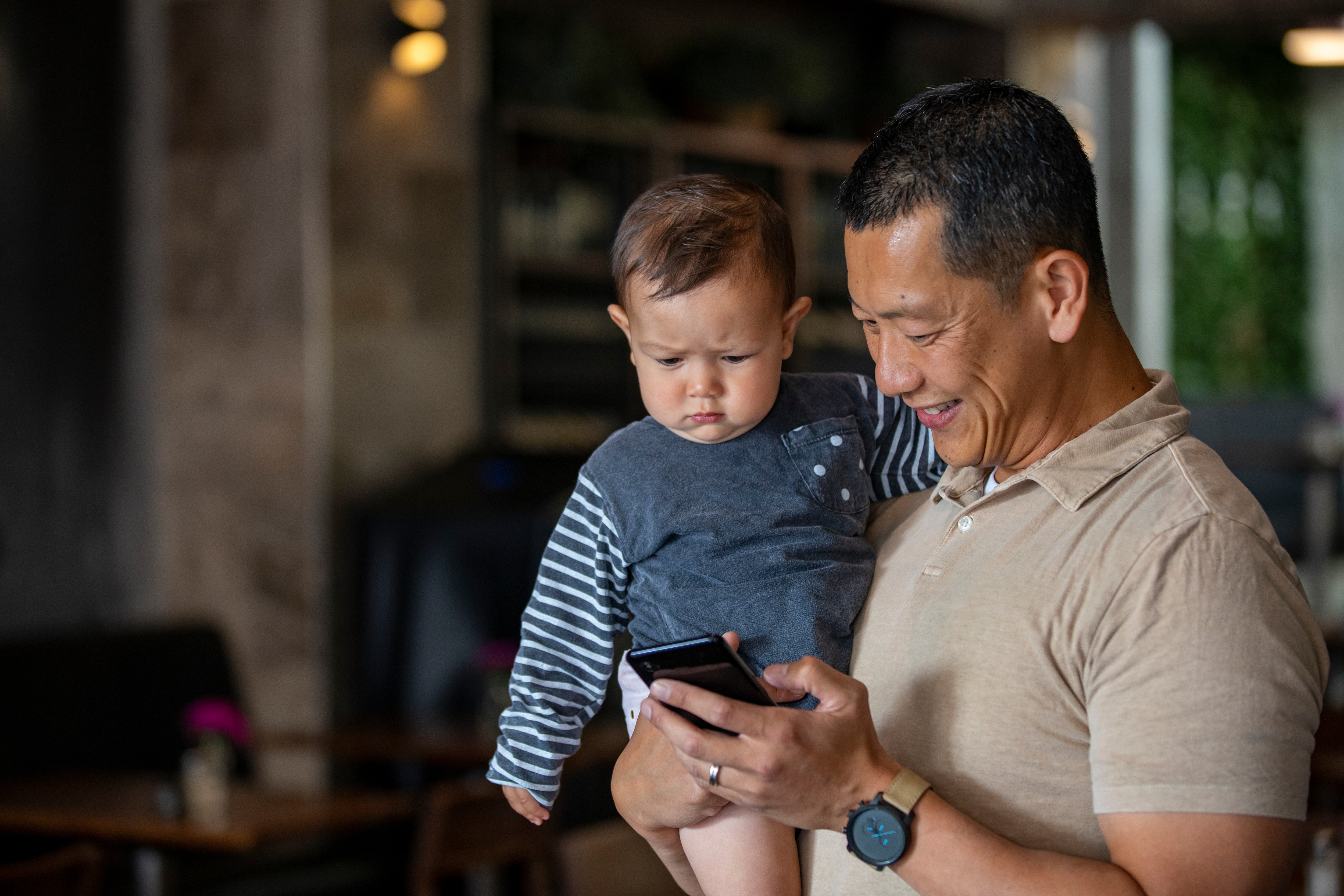 An East Asian mature male in a cafe holding his small child as he is looking at his phone