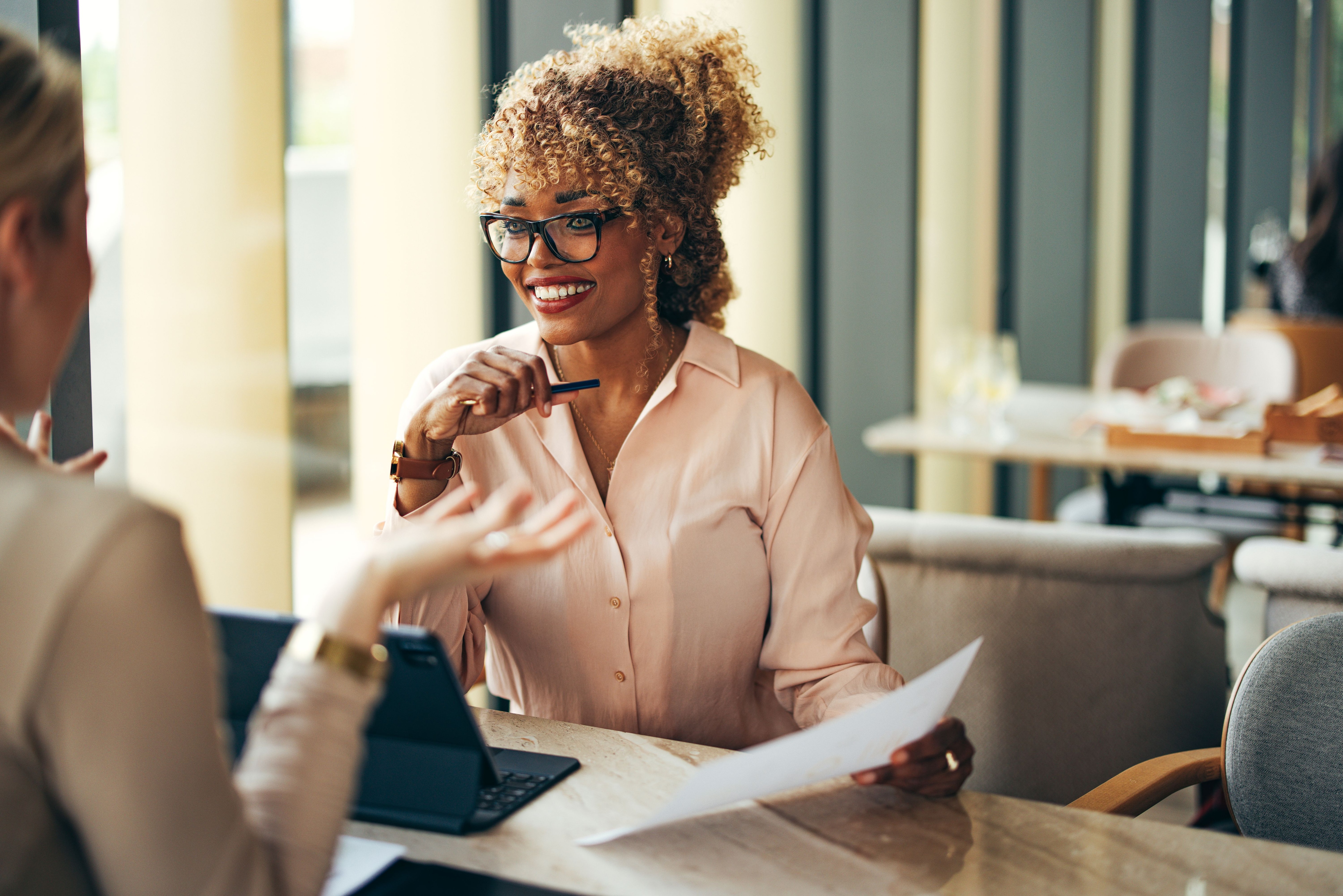 Smiling African-American businesswoman holding paper and talking with unrecognizable client or coworker while sitting together at restaurant desk with a digital tablet.