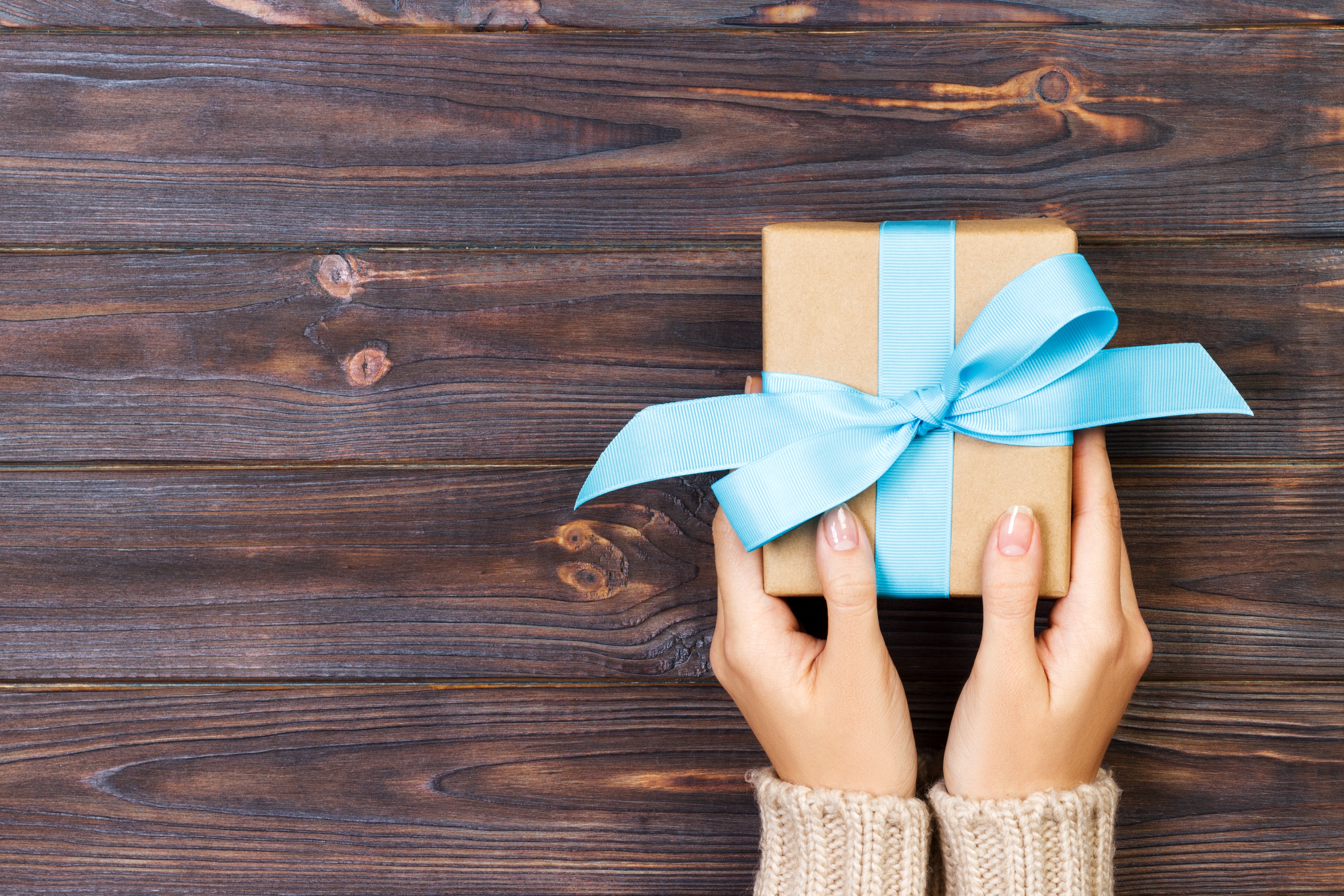 Woman hands give wrapped valentine or other holiday handmade present in paper with blue ribbon. Present box, decoration of gift on white wooden table, top view with copy space.