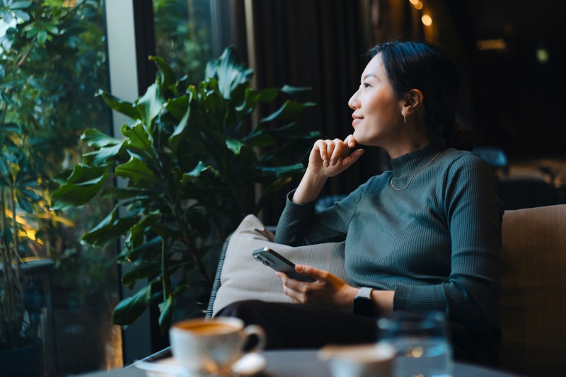 Confident young Asian woman with smartphone looking out through window while sitting in a cafe having coffee. Making a personal financial plans and investment decisions. Wealth management. Business, banking, finance and investment concept