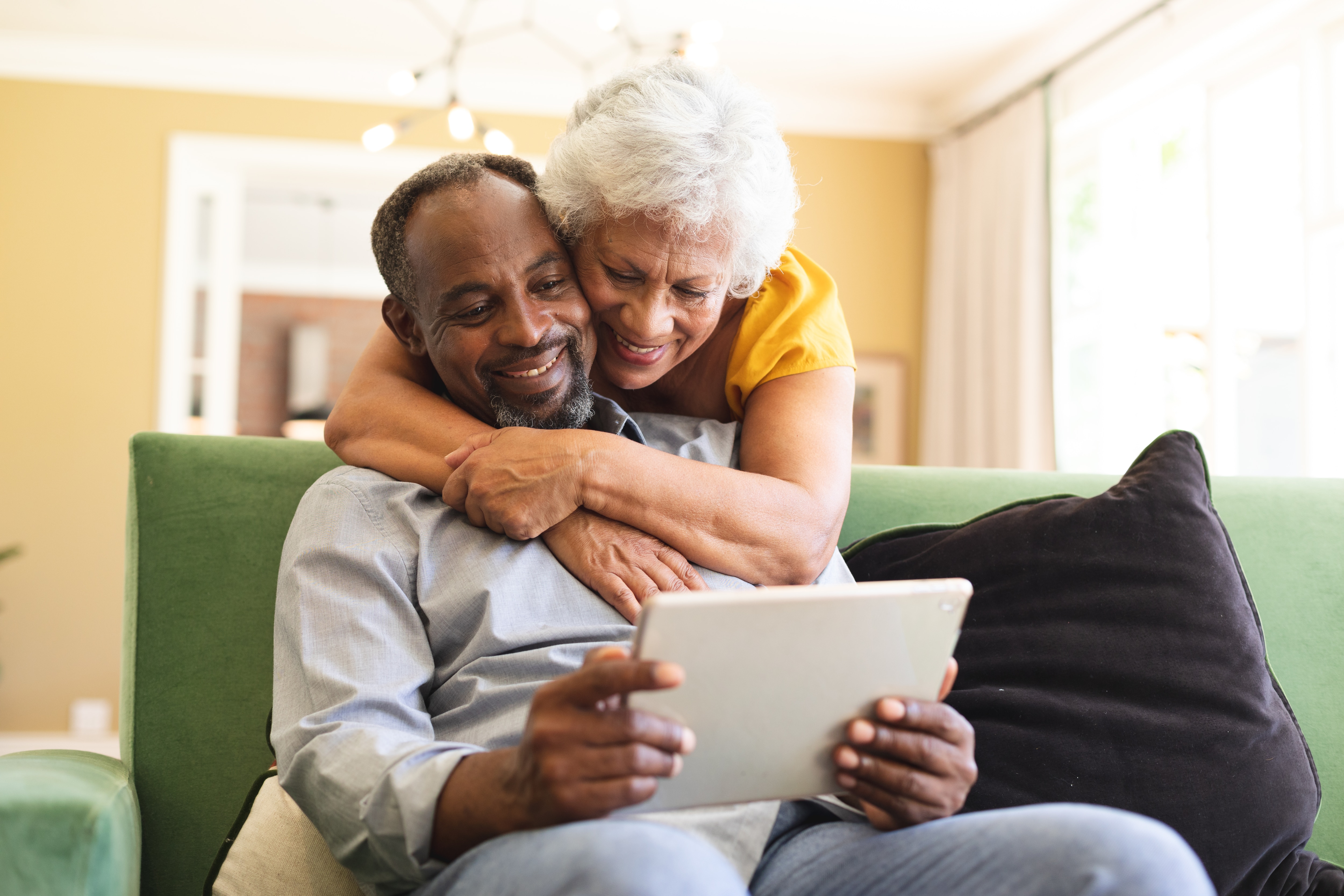 Happy senior retired African American couple at home, the man sitting on a sofa in their living room, the woman standing behind and embracing him, looking at a tablet computer together and smiling, couple isolating during coronavirus Covid19 pandemic. senior, elderly, woman, female, mixed-race, mature adult, man, male, black, people, social distancing, quarantine, self isolation, real people, isolation, distance, lock down, coronavirus, Covid19, couple, together, retirement, home, domestic life, free time, 