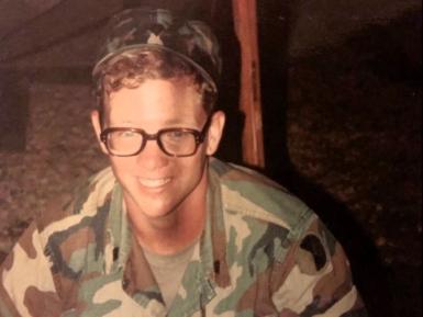 Young man in Army fatigues and glasses smiling at the camera.