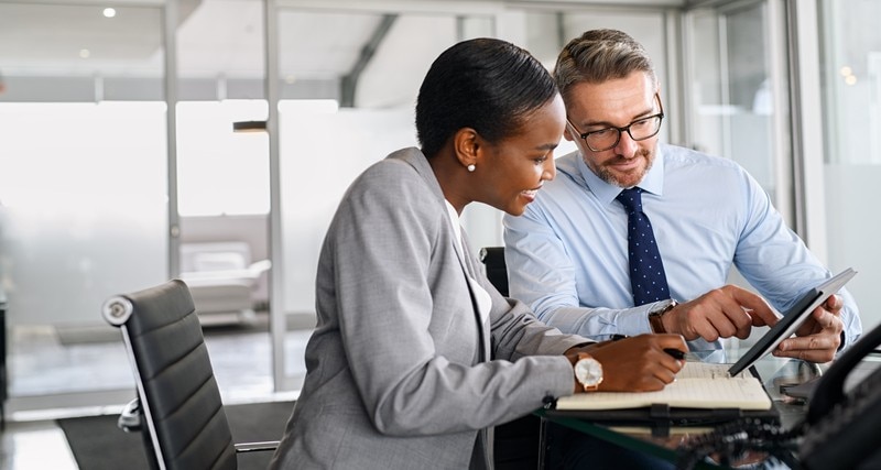Mature businessman discussing work with black manager writing notes in diary. Confident business man in formal clothing working with african american woman showing data on digital tablet. Smiling professional business people working together on new business strategy.
